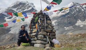 Un homme est assis devant un cairn de pierres décoré de piolets et drapeaux multicolores