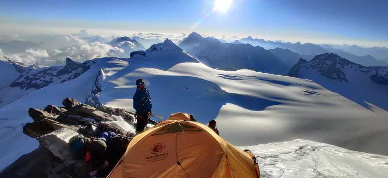 Un homme à côté d'une tente jaune admire le sommet des montagnes enneigées