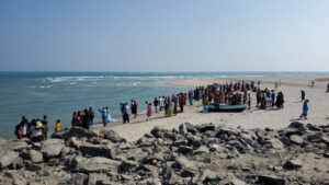 Une foule est agglutinée en bord de mer sur une plage avec au 1er plan des pierres